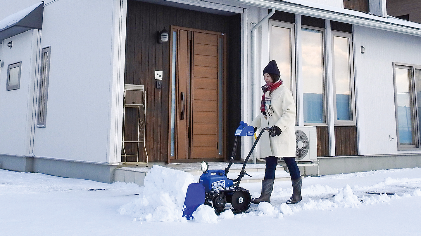 電動除雪機オ・スーノで住宅の庭の除雪をする女性。